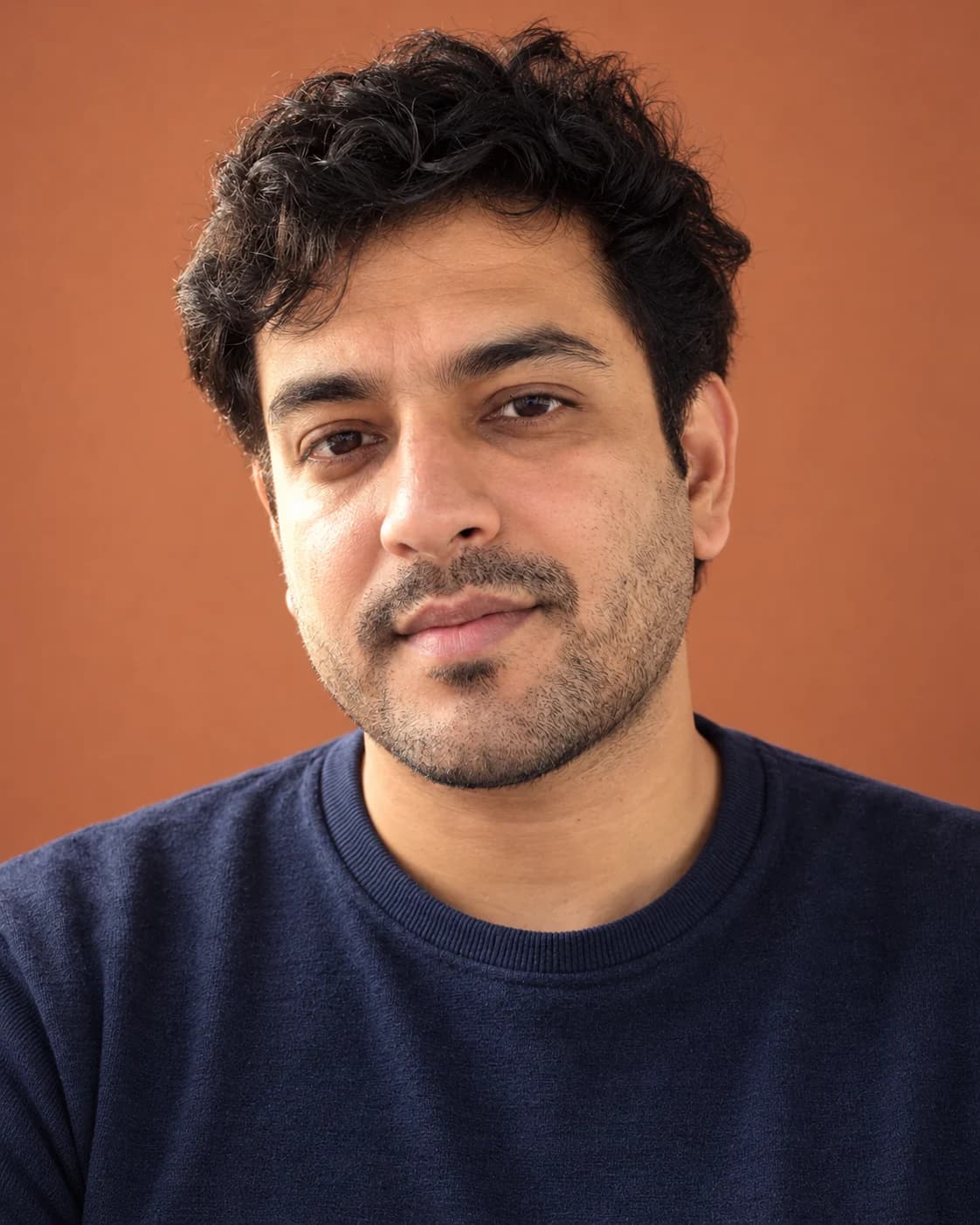 Studio headshot of a man in a navy sweater against a warm terracotta backdrop